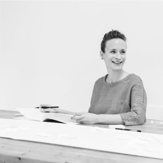 Black and white photo of a woman smiling sitting at a large wooden table with papers and a pen.