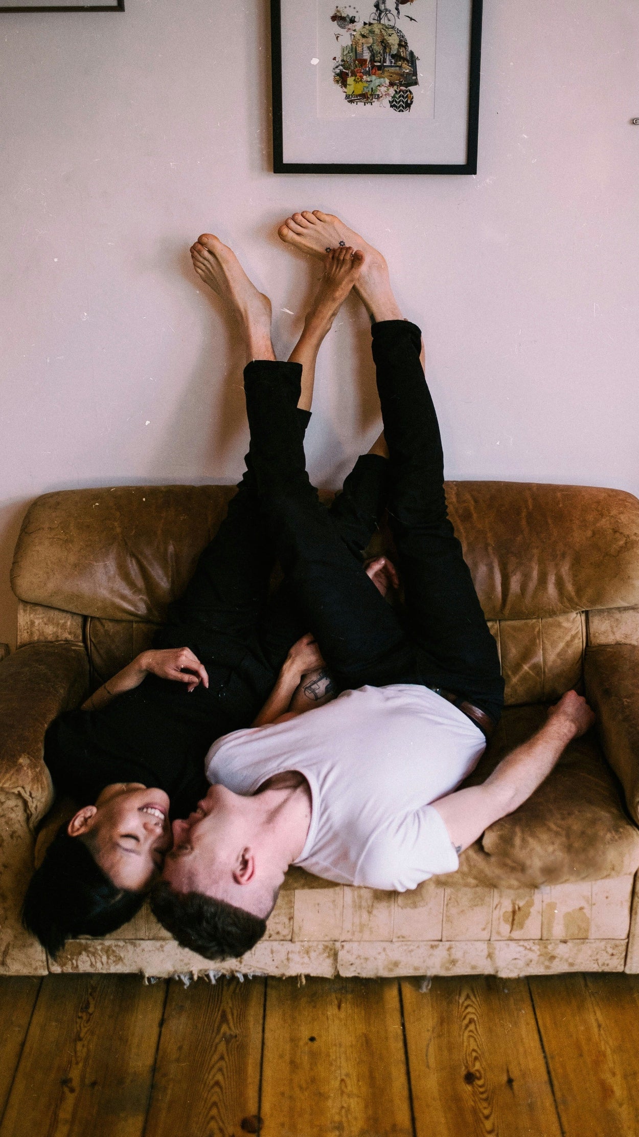 Two people smiling at each other, lying upside down on a brown couch with their feet in the air, a framed picture on the wall - photo thanks to filipp-romanovski-QzqoRzEwzeI-unsplash